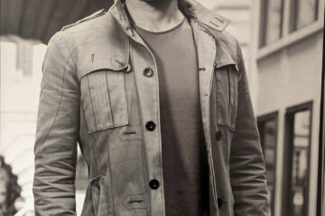Sepia-toned portrait of Carlos Simpson, a multidisciplinary artist, mentor, and creative entrepreneur, standing confidently on a city street in London. Wearing a casual jacket and jeans, he gazes directly into the camera, embodying focus, resilience, and success. The urban backdrop reinforces his connection to culture, design, and contemporary expression.