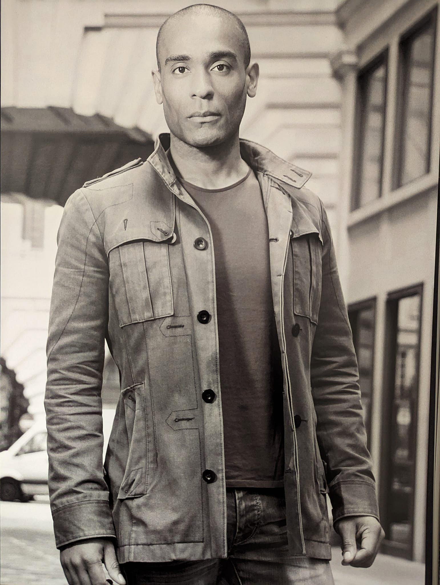 Sepia-toned portrait of Carlos Simpson, a multidisciplinary artist, mentor, and creative entrepreneur, standing confidently on a city street in London. Wearing a casual jacket and jeans, he gazes directly into the camera, embodying focus, resilience, and success. The urban backdrop reinforces his connection to culture, design, and contemporary expression.