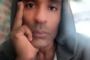 Portrait of Carlos Simpson, London-based artist, designer, and musician, wearing a grey hoodie, looking directly at the viewer.