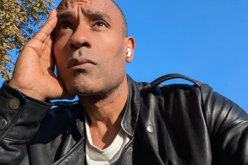 Carlos Simpson sitting outdoors under a clear blue sky, wearing a black leather jacket, reflecting thoughtfully symbolizing awareness and contemplation of humanity’s future.