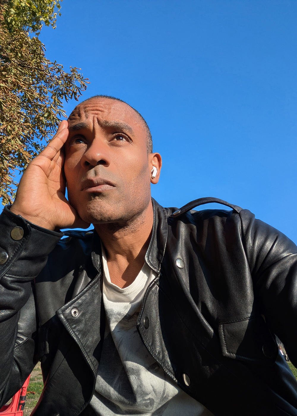 Carlos Simpson sitting outdoors under a clear blue sky, wearing a black leather jacket, reflecting thoughtfully symbolizing awareness and contemplation of humanity’s future.