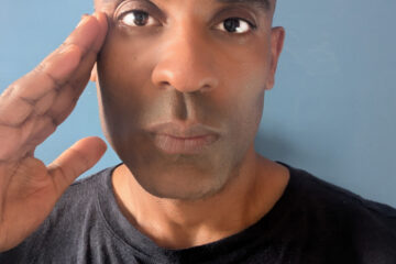 Portrait of Carlos Simpson, artist and entrepreneur, raising his right hand to his temple against a plain blue background.