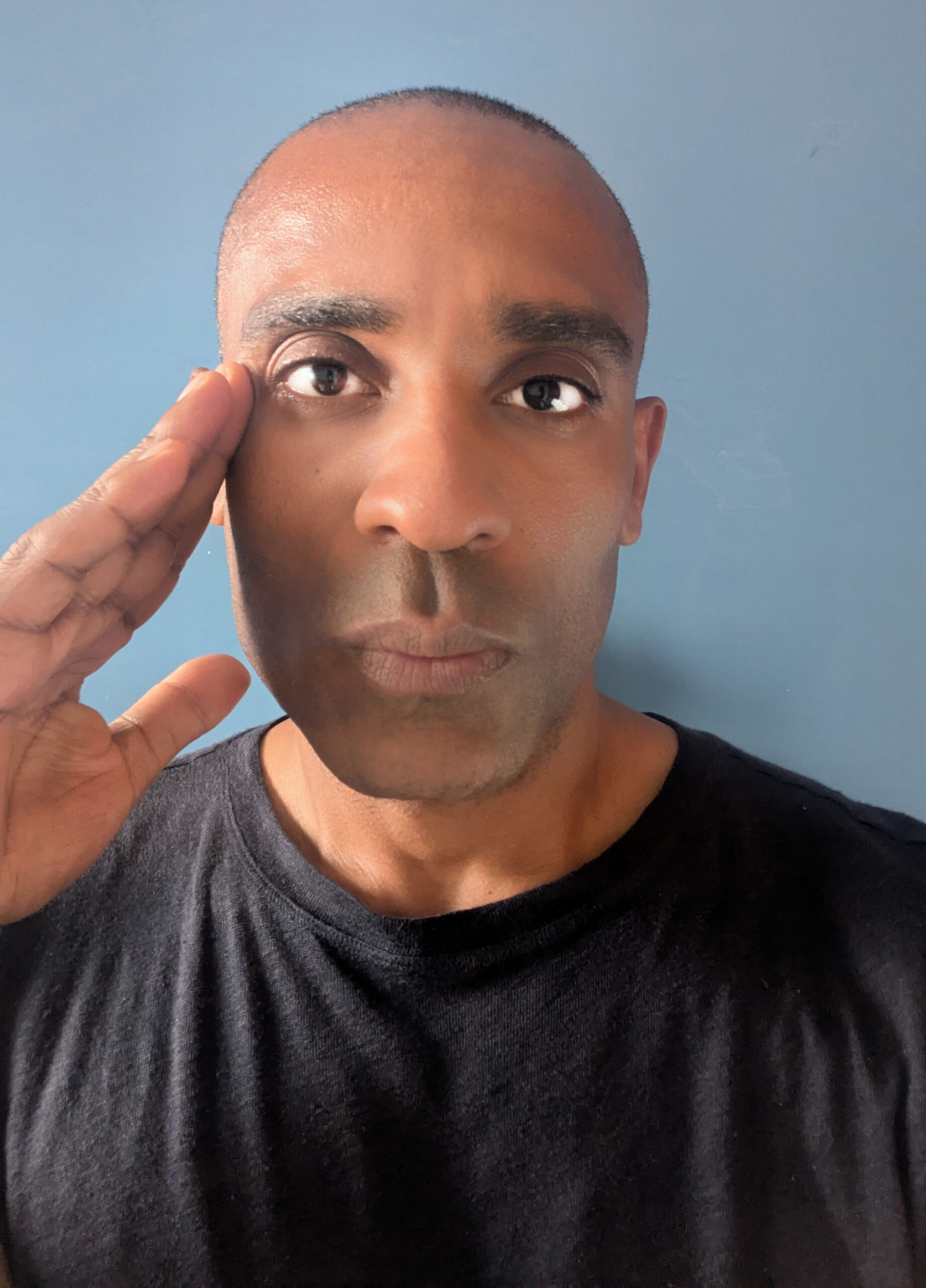 Portrait of Carlos Simpson, artist and entrepreneur, raising his right hand to his temple against a plain blue background.