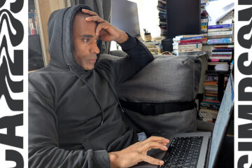 Carlos Simpson™ sits in quiet reflection at his London studio, wearing a black hoodie while working on his laptop, surrounded by stacks of books. The photo represents introspection, creativity, and mindfulness — the visual essence of his article “The Lost Frequency of Happiness,” exploring the intersection of neuroscience, emotion, and human connection.