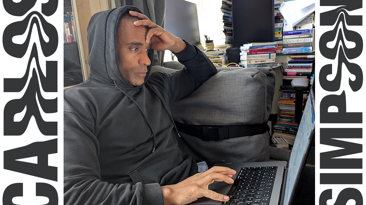 Carlos Simpson™ sits in quiet reflection at his London studio, wearing a black hoodie while working on his laptop, surrounded by stacks of books. The photo represents introspection, creativity, and mindfulness — the visual essence of his article “The Lost Frequency of Happiness,” exploring the intersection of neuroscience, emotion, and human connection.